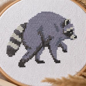 May include: A circular wooden embroidery hoop containing a white fabric panel with a cross-stitch design of a raccoon. The raccoon is depicted in shades of light purple, dark gray, and black, featuring a striped tail and a distinct facial mask.