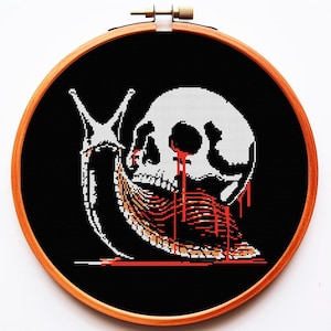 以下が含まれることがあります： 木製のフレームに入ったクロスステッチアート。赤い血が滴る白いドクロとカタツムリの体を組み合わせたデザイン。背景は黒色です。