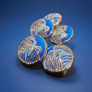 Op de afbeelding: Vijf blauwe en gouden decoratieve kastknoppen. Elke knop heeft een gouden rand en een blauw emaille ontwerp met gouden accenten. De ovaalvormige knoppen zijn gerangschikt op een blauw oppervlak.