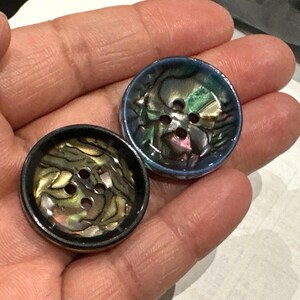 May include: Two round buttons with iridescent, shell-like centres and four holes. One button has a black rim, the other has a blue rim. The buttons are set against a neutral background.