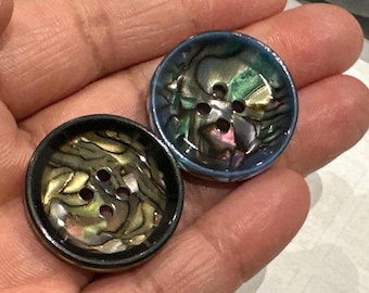 May include: Two round buttons with iridescent, shell-like centers and four holes. One button has a black rim, the other has a blue rim. The buttons are set against a neutral background.