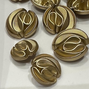 Może przedstawiać: Zbiór złotych metalowych guzików z ozdobnym, spiralnym wzorem. Guziki są okrągłe i mają wypukłą, fakturowaną powierzchnię.