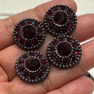 Op de afbeelding: Vier ronde, donkergrijze knopen met een centraal, groot, rood juweel omringd door kleinere rode juwelen. De knopen hebben een decoratieve, kralenrand. De knopen worden in een hand gehouden.