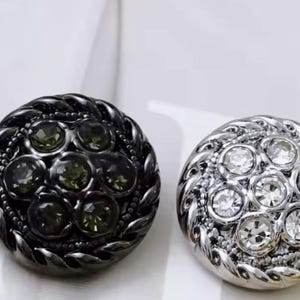 May include: Two decorative buttons. One is dark gray with olive green gemstones, the other is silver with clear gemstones. Both buttons have a braided edge and are round in shape.