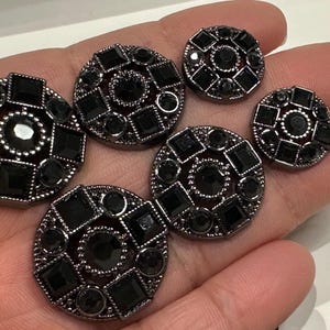 Puede incluir: Una colección de botones decorativos negros con un acabado de metal de cañón oscuro. Los botones presentan una joya central rodeada de gemas negras cuadradas y redondas. Los botones varían en tamaño y están dispuestos sobre un fondo claro.
