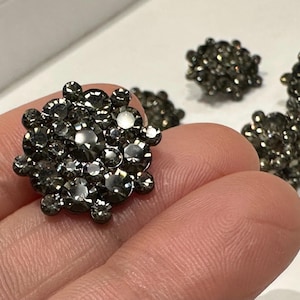 Puede incluir: Primer plano de cuatro botones de pedrería gris. Los botones son redondos y tienen un diseño en relieve. Son perfectos para añadir un toque de brillo a cualquier proyecto.
