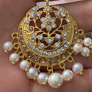 Może przedstawiać: Złoty, ozdobny kolczyk o okrągłym kształcie. Kolczyk posiada wzór filigranowy, zdobiony przezroczystymi i brązowymi kryształami. Białe perły w kształcie gruszki zwisają z dołu, dodając klasycznego charakteru tej biżuterii.