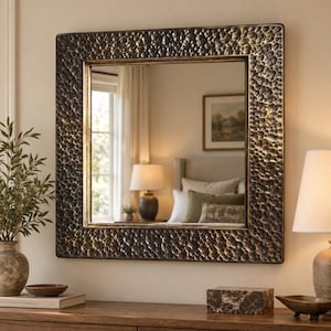 May include: A square wall mirror featuring a wide, dark bronze frame with a heavily hammered, textured surface. The reflective glass surface shows a room interior with a lamp, a bed, and a window, creating a deep, dimensional appearance.