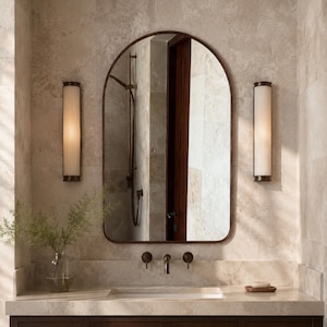 Peut inclure: Un miroir mural en arche avec un cadre fin en métal sombre, centré au-dessus d'un meuble-lavabo. Le miroir reflète une pomme de douche et une porte. De chaque côté, deux appliques murales cylindriques en verre strié avec des bases en métal sombre.