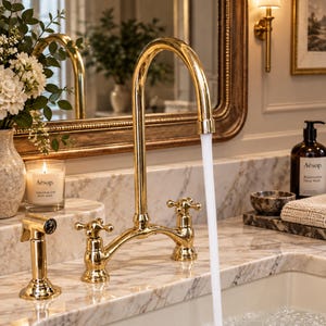 May include: A polished gold bridge tap with two cross-handle knobs and a side sprayer, mounted on a white marble countertop. A steady stream of water flows from the high-arc spout into a sink, with a candle and soap bottle nearby.