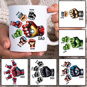 Op de afbeelding: Een witte keramische mok met een ontwerp van superhelden vuisten rond een grotere vuist in het midden. De grotere vuist is die van Iron Man. De mok heeft het woord "DAD" en verschillende namen rond het ontwerp.
