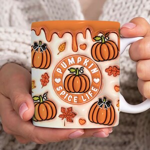 May include: White ceramic mug with a pumpkin spice life design. The mug has a dripping orange glaze and features multiple illustrations of pumpkins with black stems and leaves. The text "Pumpkin Spice Life" is written in a black circle with a heart on each side.