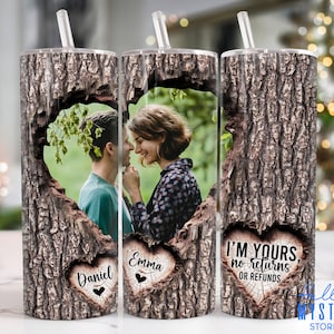 May include: Two personalized stainless steel tumblers with a tree bark design and a heart-shaped cutout. The cutout features a photo of a couple and the names "Daniel" and "Emma" are inscribed in the heart. The third tumbler has the text "I'm Yours, No Returns or Refunds".