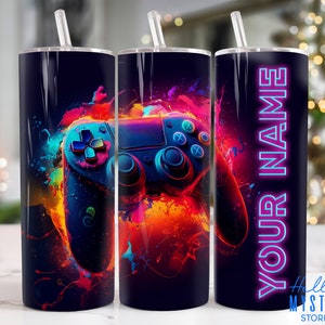 May include: A black and colorful tumbler with a video game controller design. The controller is surrounded by splashes of paint in shades of red, orange, yellow, blue, and green. The tumbler has a clear lid and a straw.