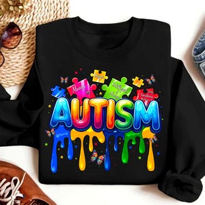 Puede incluir: Sudadera negra con un gr&aacute;fico colorido de piezas de rompecabezas que deletrean "AUTISMO". Las piezas del rompecabezas tienen texto que dice "Siempre", "Inteligente y siempre", y "A veces rutina".