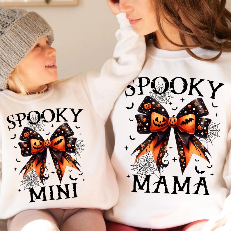May include: White long-sleeved shirts with a black and orange bow with a pumpkin design. The shirt on the left says "Spooky Mini" and the shirt on the right says "Spooky Mama".