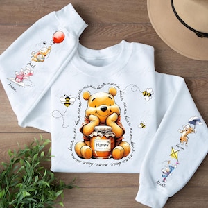 Può includere: Felpa bianca con un design di Winnie the Pooh. Pooh tiene un barattolo di miele e è circondato da api. Il testo "mana bear mama bear mama bear mama bear mama bear nana bear mama bear mama" è scritto intorno a Pooh. La felpa ha altri personaggi Disney sulle maniche, tra cui Tigro, Maialino e Ih-Oh.