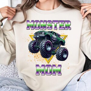 May include: A white sweatshirt with the text "Monster Mom" in purple and green letters. A green monster truck with a skull on the hood is in the center of the sweatshirt.