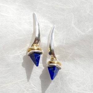 May include: A pair of silver earrings with blue gemstone accents and gold accents. The earrings are shaped like a long, thin point.