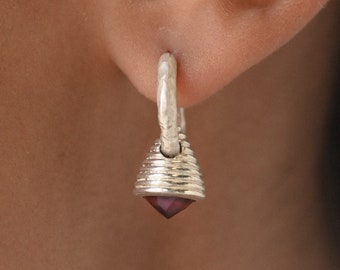 May include: Silver hoop earrings with a small, faceted, dark purple gemstone hanging from the bottom. The earrings are worn in a person's ear.