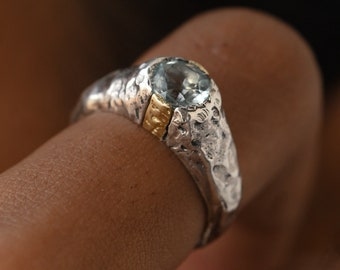 May include: A silver ring with a textured finish and a light blue gemstone set in a gold band.