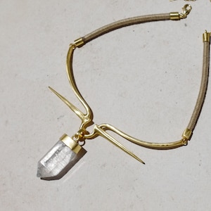 May include: A gold necklace with a clear crystal pendant and a brown cord. The necklace has a unique design with a curved bar and a pointed spike.