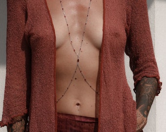 May include: A red, sheer, open-front cardigan with a silver chain body jewelry featuring red gemstones. The jewelry is worn over a dark red, tie-dye skirt.