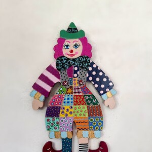 May include: A colorful wooden clown doll with a patchwork dress, green hat, pink hair, and red shoes. The doll is painted with various patterns and colors.