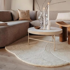 Könnte beinhalten: Ein runder, cremefarbener Teppich mit einem strukturierten, konzentrischen Wellenmuster. Der Teppich liegt auf einem hellen Holzboden unter einem runden Couchtisch mit heller Tischplatte und dünnen Metallbeinen, auf dem zwei weiße Kerzen stehen.