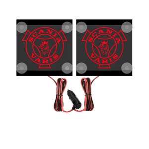 Może przedstawiać: Dwa czarne kwadratowe naklejki na szyby samochodowe z czerwonym logo Scania Vabis. Naklejki są podświetlane czerwonymi diodami LED i są dostarczane z adapterem do zapalniczki.