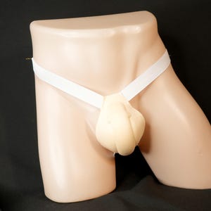 May include: A beige prosthetic device designed to simulate male genitalia. It features a soft, rounded form held in place by a white elastic waistband. The device is displayed on a mannequin against a dark background.