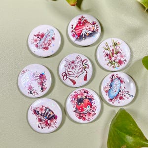 Könnte beinhalten: Eine Sammlung von acht weißen, runden Knöpfen mit japanisch inspirierten Designs. Jeder Knopf zeigt eine andere Illustration, darunter Kirschblüten, eine Fuchmaske, eine Feder und eine Berglandschaft. Die Knöpfe haben einen Durchmesser von etwa 2,5 cm.
