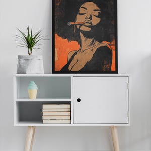 May include: A black and white illustration of a woman with a large afro, wearing a black top and smoking a cigar. The illustration is framed in black and hanging on a white wall above a white cabinet with wooden legs.