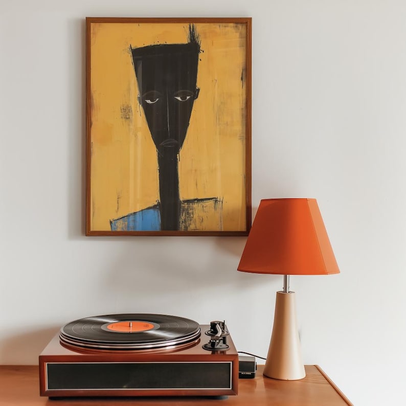 May include: A framed wall art print featuring a stylised black figure against a yellow background, positioned above a dark wood record player with a black vinyl disc and a table lamp with a conical orange shade and a beige base.