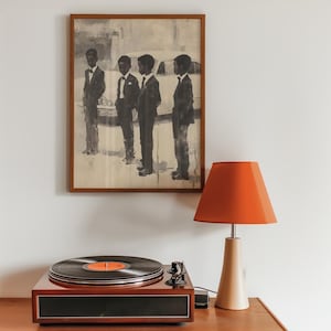 May include: A framed monochromatic art print depicting four figures in suits standing before a car, positioned above a dark wood record player with a black vinyl disc and a table lamp featuring a vibrant orange conical shade and a beige base.