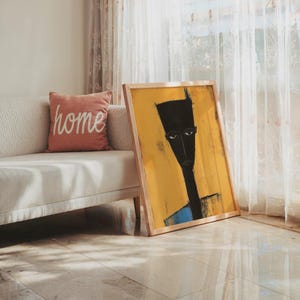 May include: A framed rectangular wall art print featuring a stylised, elongated black figure against a vibrant yellow background. The figure has a flat-topped head, almond-shaped eyes, and a thin neck, set within a light-coloured wooden frame.