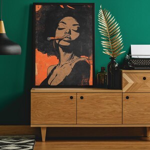 May include: A black and white illustration of a woman with an afro, wearing a black top and smoking a cigar. The illustration is set against an orange background and framed in a black frame. The artwork is displayed on a wooden cabinet with a typewriter and a gold leaf branch.