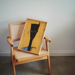 May include: A rectangular framed print featuring a stylised black figure with an elongated head and torso set against a solid yellow background. The figure has a small blue section at the bottom. The artwork is displayed on a light wood chair.