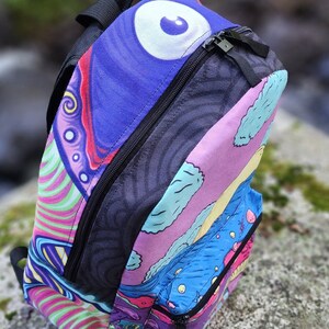 May include: A colorful psychedelic backpack with a large eye design on the front. The backpack has a black zipper and a black strap.