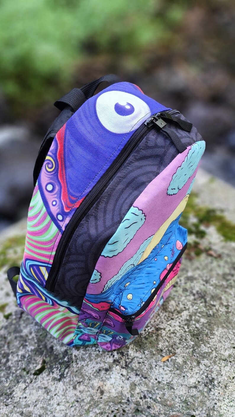 May include: A colorful psychedelic backpack with a large eye design on the front. The backpack has a black zipper and a black strap.