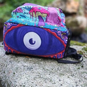 May include: A psychedelic backpack with a large eye design in shades of purple, blue, and white. The backpack has a colorful, abstract pattern with mushrooms and other whimsical elements.