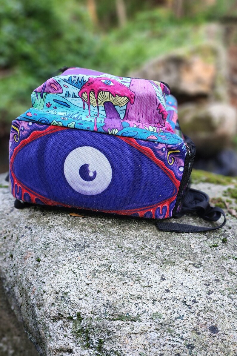 May include: A psychedelic backpack with a large eye design in shades of purple, blue, and white. The backpack has a colorful, abstract pattern with mushrooms and other whimsical elements.