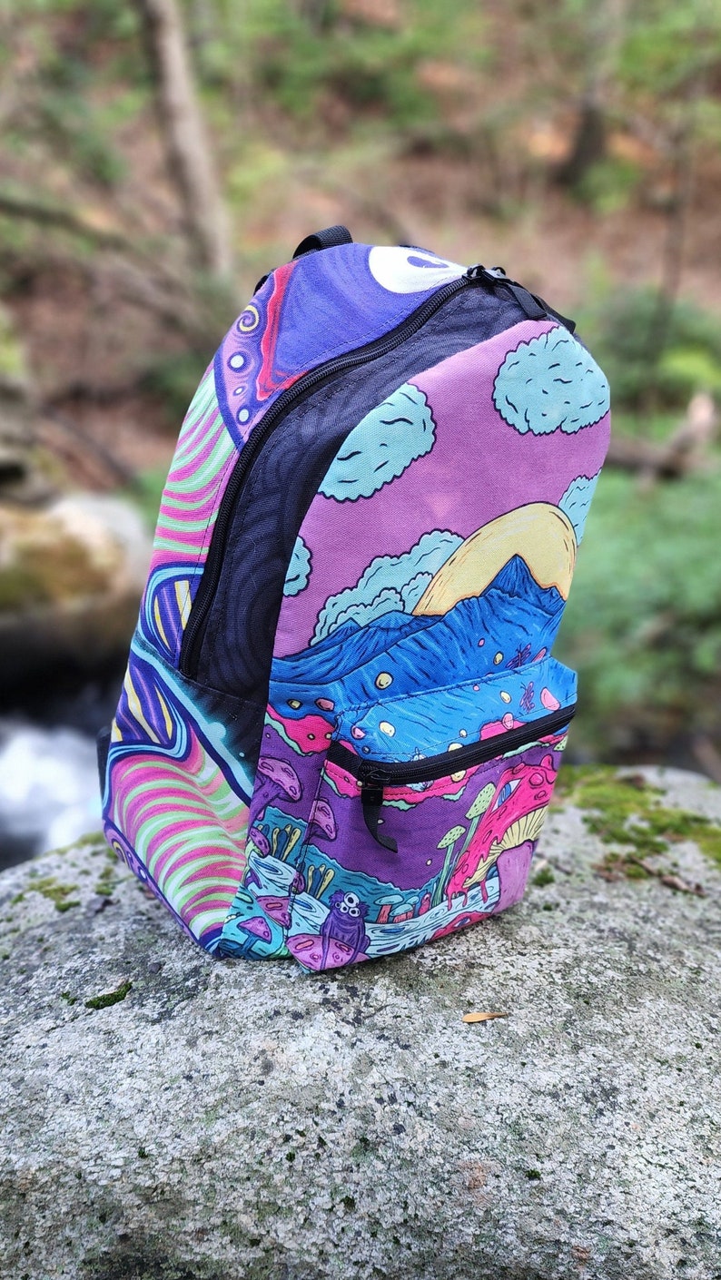 May include: A psychedelic backpack with a purple, pink, blue, and green design. The backpack features a mountain scene with a sun and clouds, as well as mushrooms and a three-eyed creature.