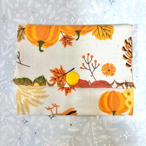 May include: A rectangular fabric pouch featuring a seasonal print of orange pumpkins, yellow and brown autumn leaves, and sprigs of berries on a cream background. The flap is secured with a single yellow snap button and finished with brown decorative stitching.