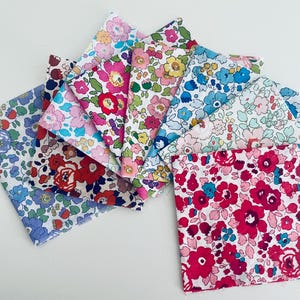 May include: A collection of square fabric swatches arranged in a fan shape. Each piece features a distinct floral pattern with vibrant colours including shades of pink, red, blue, yellow, and green on white backgrounds, showcasing various botanical motifs.