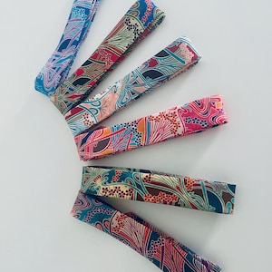 Peut inclure: Six bandeaux en tissu disposés en éventail, chacun présentant une palette de couleurs distincte incluant des tons de bleu, rouge, rose, sarcelle et marron. Le tissu affiche un motif répétitif de fleurs stylisées et de lignes organiques.