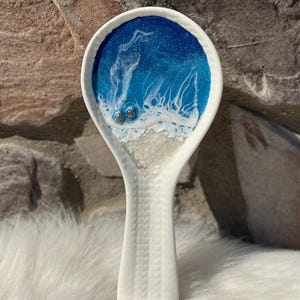 May include: White ceramic spoon rest with a blue and white ocean-themed design. The spoon rest features a textured handle and a circular top with a resin ocean scene, complete with tiny turtle accents.