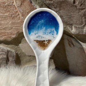May include: White ceramic spoon rest with a beach scene design. The spoon rest features a blue and white ocean design with a sandy beach and a starfish. The handle has a quilted pattern.