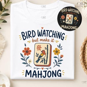 May include: A white t-shirt featuring a graphic print with the text BIRD WATCHING but make it MAHJONG. The design includes a central mahjong tile depicting a bird on a bamboo branch, surrounded by orange and yellow flowers and small red hearts.