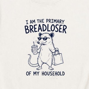 May include: An off-white short-sleeve t-shirt featuring a dark blue graphic of an opossum wearing sunglasses, holding a drink and a shopping bag. The text reads I AM THE PRIMARY BREADLOSER OF MY HOUSEHOLD in a dark blue sans-serif font.